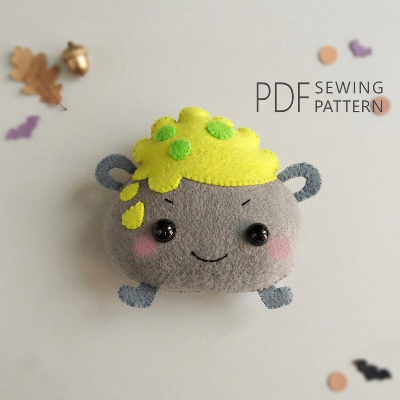 May include: A gray felt plush cauldron with two handles, a smiling face, and pink cheeks. The top features yellow bubbling liquid with green spots. The text PDF SEWING PATTERN is visible in the upper right corner, accompanied by small felt shapes.