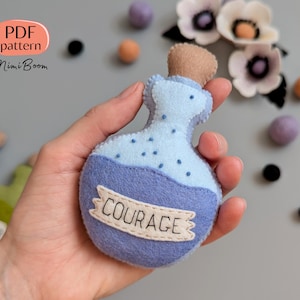 Puede incluir: Una botella de poción de fieltro azul con tapón de corcho y la palabra "COURAGE" en una pancarta. La imagen también incluye flores y formas de fieltro. El texto "PDF pattern MimiBoom" es visible en la esquina superior izquierda.