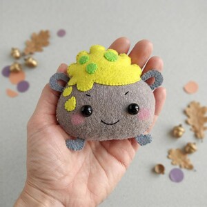 May include: A gray felt cauldron plushie with a yellow bubbling potion top featuring green spots. The cauldron has two small handles, black bead eyes, a stitched smile, and pink blush marks on its cheeks, crafted from soft felt material.