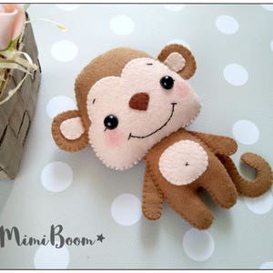 May include: A brown felt monkey toy with a pink belly and a stitched smile. The monkey has black eyes and a brown nose.