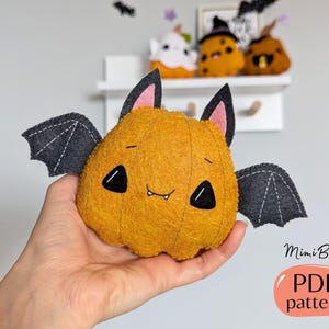 May include: A plush, orange pumpkin-shaped bat with black eyes, a small smile, and grey wings. The bat has black ears with pink inner ears. The image also includes other plush pumpkins on a shelf and the text "MimiBoom PDF pattern".