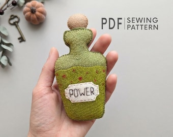 Pode incluir: Um ornamento de frasco de poção em feltro verde-oliva com uma rolha redonda bege. O frasco apresenta um rótulo branco com a palavra POWER bordada em linha preta e pequenos detalhes de estrelas vermelhas. O texto PDF SEWING PATTERN é visível.