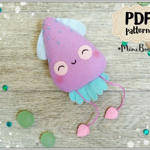 Puede incluir: Un juguete de pulpo de fieltro hecho a mano en tonos morado, azul claro y rosa. El pulpo tiene una cara sonriente, detalles de cuentas azules y tentáculos rosas colgantes. La imagen incluye el texto "PDF pattern" y el logotipo de Mimi Boom.