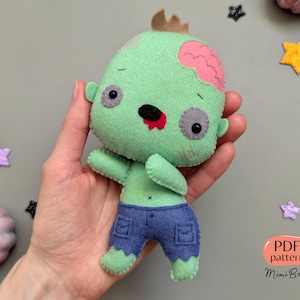 Peut inclure: Peluche zombie en feutre vert avec un cerveau rose, une bouche rouge et des yeux noirs. Le jouet porte un jean bleu et est tenu par une main. Le texte "PDF pattern Mimi Boom" est dans le coin inférieur droit de l'image.