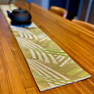 May include: A decorative table runner with a gold and green leaf pattern. A black teapot sits on the runner. The runner is rectangular and made of fabric. The table is made of wood.