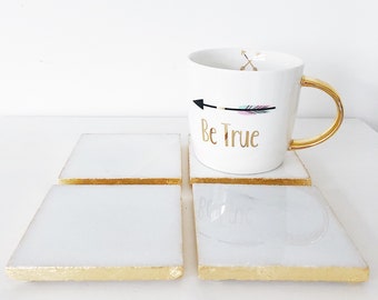 May include: Four white square coasters with gold trim and a white mug with a gold handle. The mug has a black and pink arrow with the text "Be True" in gold lettering.