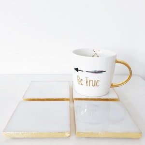 May include: Four white square coasters with gold trim and a white mug with a gold handle. The mug has a black and pink arrow with the text "Be True" in gold lettering.