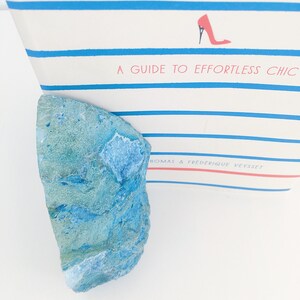 May include: A blue and green agate stone with a rough, natural texture. The stone is sitting on a white surface.