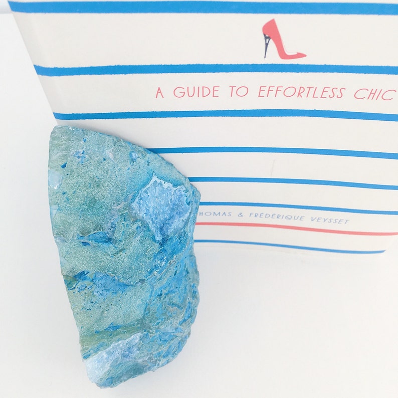 May include: A blue and green agate stone with a rough, natural texture. The stone is sitting on a white surface.