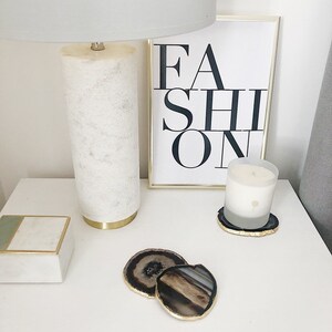 May include: A white lamp with a gold base sits on a white nightstand next to a framed print that says "FASHION". A white candle sits on a gold and black agate coaster. Two more agate coasters are on the nightstand.