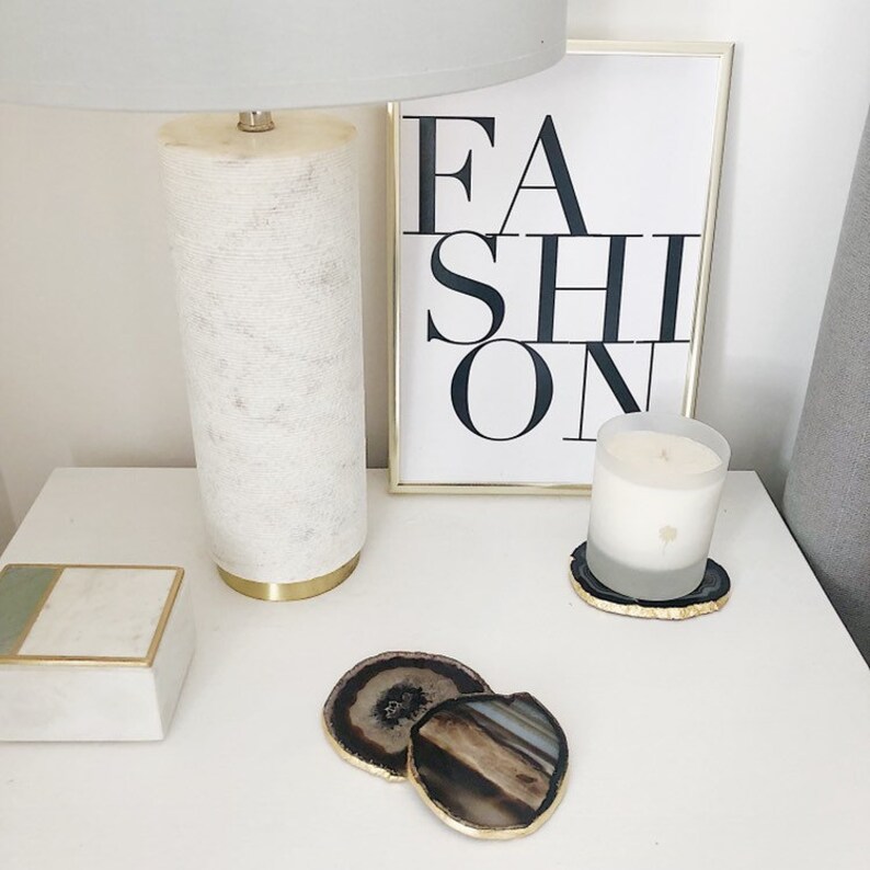 May include: A white lamp with a gold base sits on a white nightstand next to a framed print that says "FASHION". A white candle sits on a gold and black agate coaster. Two more agate coasters are on the nightstand.