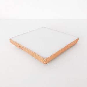 May include: Square white ceramic coaster with a copper-colored edge. The coaster is a simple, modern design, perfect for protecting surfaces from drink condensation. The copper edge adds a touch of elegance.