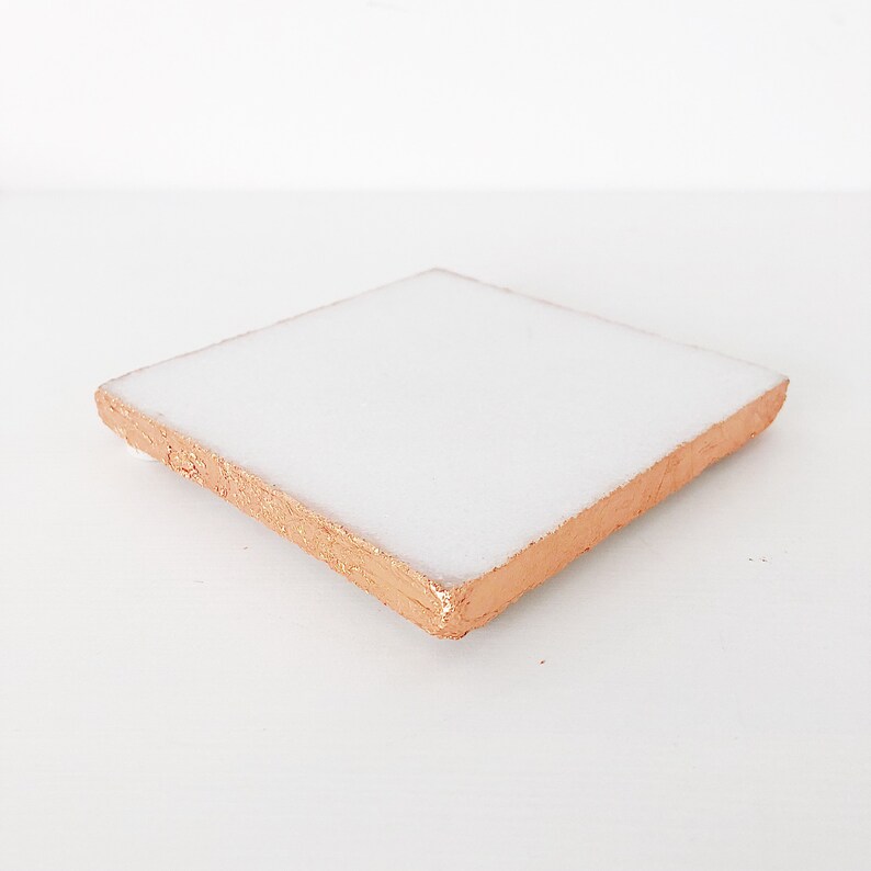May include: Square white ceramic coaster with a copper-colored edge. The coaster is a simple, modern design, perfect for protecting surfaces from drink condensation. The copper edge adds a touch of elegance.