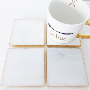 May include: Four white square coasters with gold trim. The coasters are made of marble and have a subtle speckled pattern. A white mug with gold trim and the text "Be True" is sitting on top of two of the coasters.
