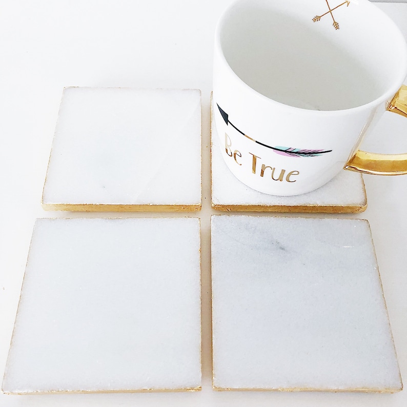 May include: Four white square coasters with gold trim. The coasters are made of marble and have a subtle speckled pattern. A white mug with gold trim and the text "Be True" is sitting on top of two of the coasters.
