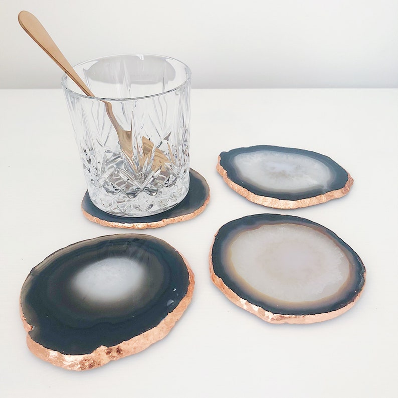 May include: Four agate coasters with a copper rim. The coasters are round and have a black and white pattern. The coasters are on a white surface.
