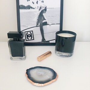 May include: A black and white photo of a woman holding a surfboard with the word "Chanel" printed on it. A black candle, a black perfume bottle, and a rose gold lipstick are on a white surface.
