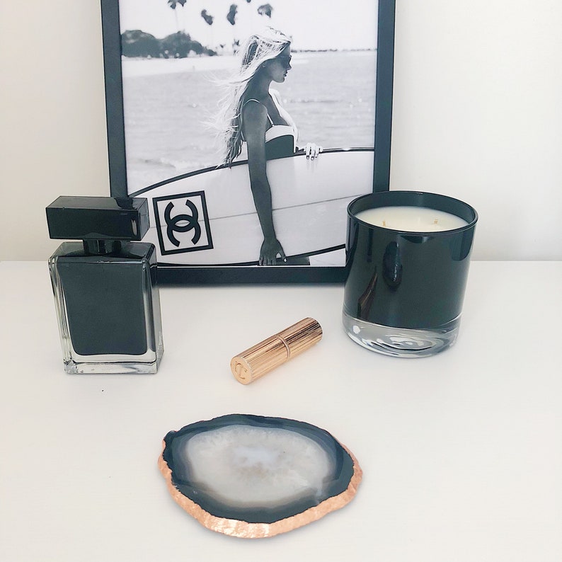May include: A black and white photo of a woman holding a surfboard with the word "Chanel" printed on it. A black candle, a black perfume bottle, and a rose gold lipstick are on a white surface.