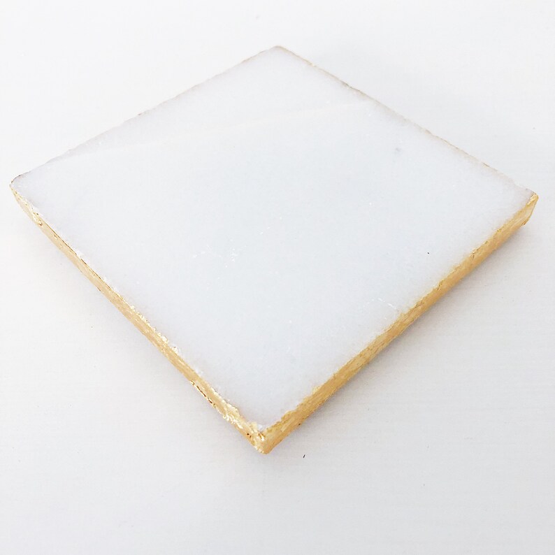 May include: A white square tile with a gold border. The tile is smooth and has a slightly glossy finish.