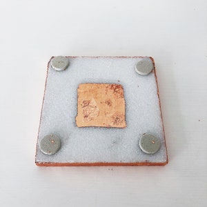 May include: Square coaster with a light brown border, a light gray center, and four small, round, gray feet. A piece of textured, light brown material is adhered to the center of the coaster.