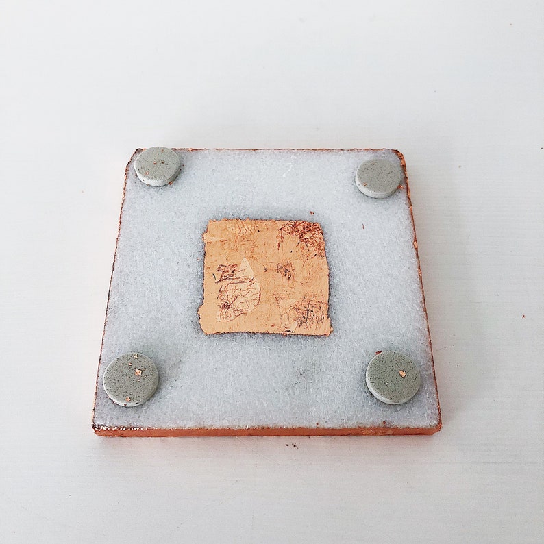 May include: Square coaster with a light brown border, a light gray center, and four small, round, gray feet. A piece of textured, light brown material is adhered to the center of the coaster.