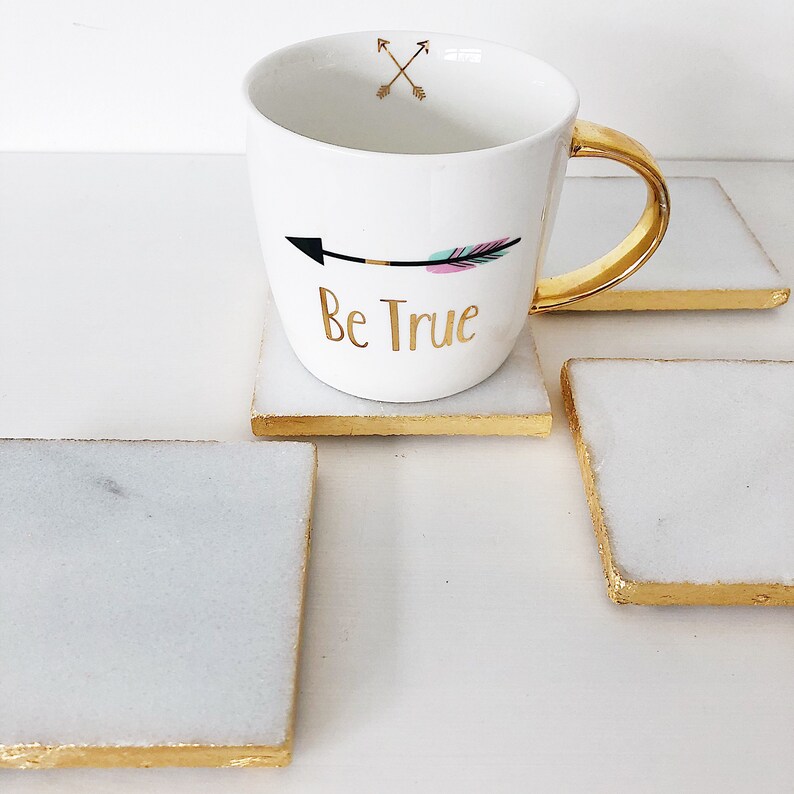 May include: White ceramic mug with a gold handle and gold rim. The mug has a black arrow with pink and blue feathers and the text "Be True" in gold lettering. The mug is sitting on a white square coaster with a gold rim.