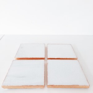 May include: Four square white marble coasters with copper-colored edges are arranged on a white surface. The coasters have a polished, reflective surface, and the copper edges add a touch of warmth and contrast to the design. These coasters are ideal for protecting surfaces from drink rings.