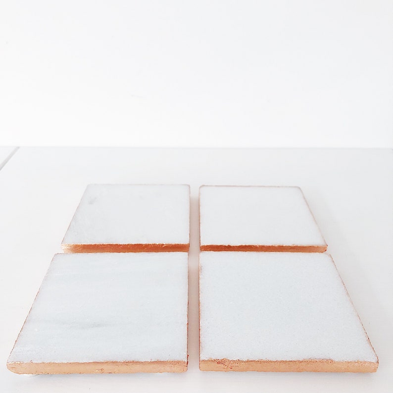 May include: Four square white marble coasters with copper-colored edges are arranged on a white surface. The coasters have a polished, reflective surface, and the copper edges add a touch of warmth and contrast to the design. These coasters are ideal for protecting surfaces from drink rings.