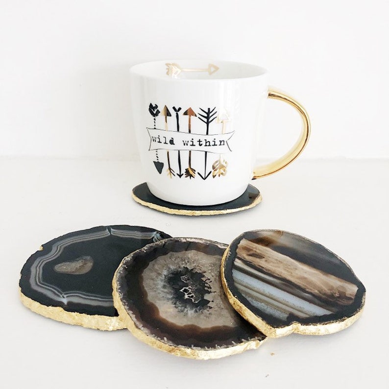 May include: Four agate coasters with gold trim. The coasters are in a stack with the top coaster showing a black and white agate pattern. The bottom coaster shows a brown and white agate pattern. A white mug with gold trim and black arrows and the text "wild within" is sitting on a coaster.