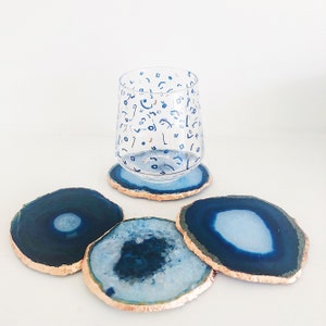 May include: A clear glass with a blue and white pattern sits on a blue agate coaster with a gold edge. Three additional blue agate coasters with gold edges are displayed. The coasters are a decorative home accessory.