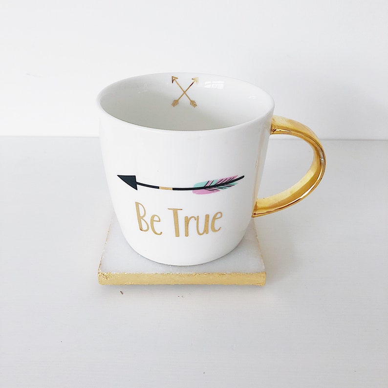 May include: White ceramic mug with a gold handle and a gold arrow with feathers design. The mug says "Be True" in gold lettering. The mug is sitting on a white square coaster with a gold trim.