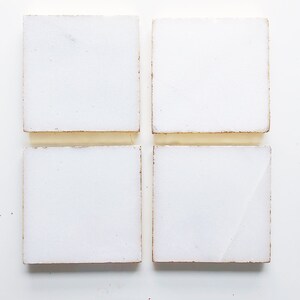 May include: Four white square tiles with a slightly rough texture and a subtle gold border.