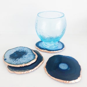 May include: Blue agate coasters with a metallic gold edge are displayed with a blue glass. The coasters have a natural, organic shape and varying shades of blue. The glass has a textured pattern.
