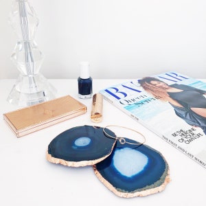 May include: A still-life arrangement featuring two blue agate coasters with gold edges, a gold compact, lipstick, nail polish, a bracelet, and a magazine. The magazine cover features a woman and the text "Queen".