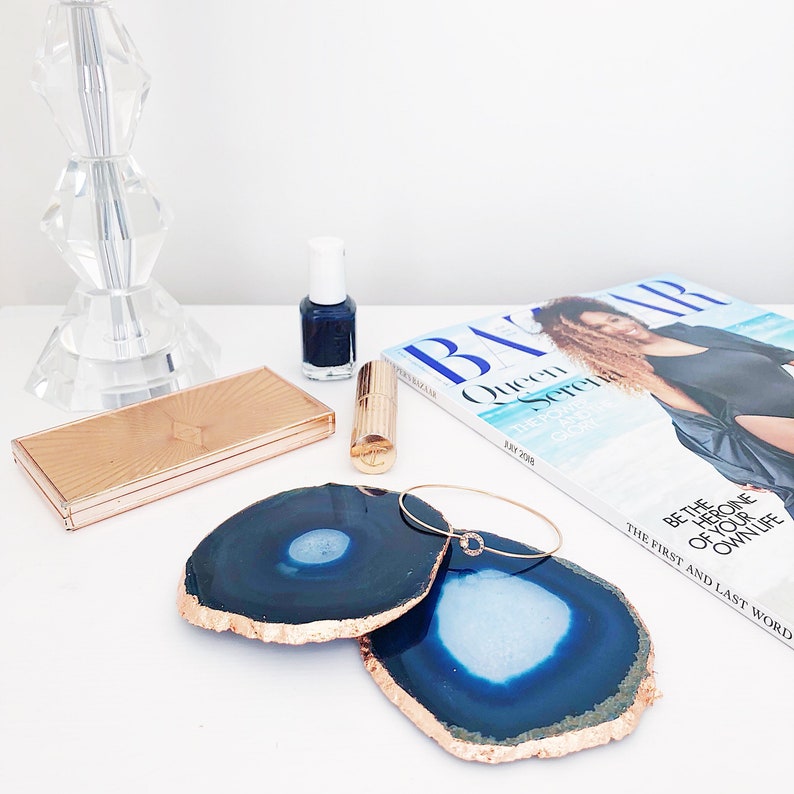 May include: A still-life arrangement featuring two blue agate coasters with gold edges, a gold compact, lipstick, nail polish, a bracelet, and a magazine. The magazine cover features a woman and the text "Queen".