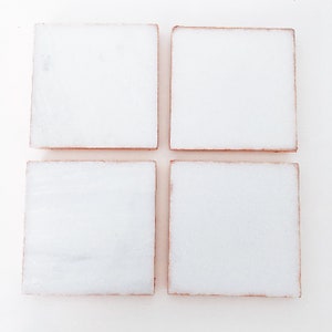 May include: Four square white coasters with a light brown edge are arranged in a grid pattern. The coasters appear to be made of a stone material, and are set against a white background.