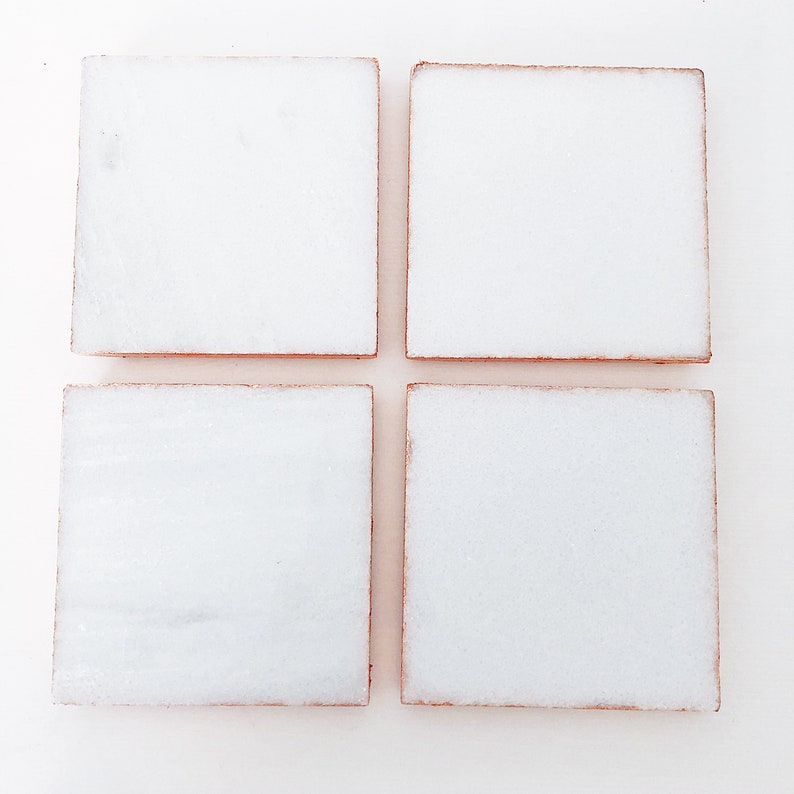 May include: Four square white coasters with a light brown edge are arranged in a grid pattern. The coasters appear to be made of a stone material, and are set against a white background.