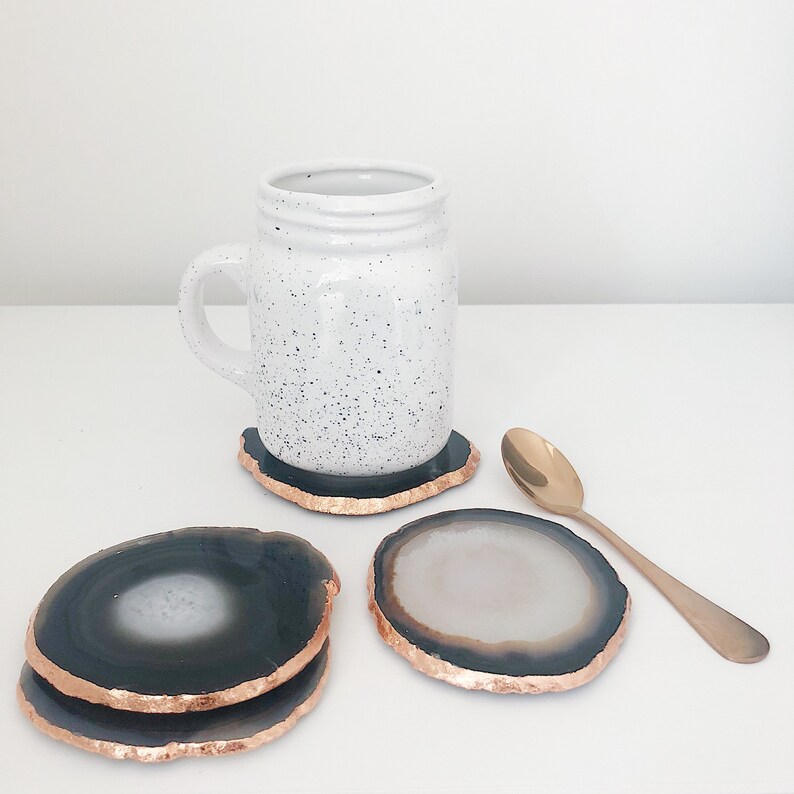 May include: Four agate coasters with gold trim. The coasters are round and have a dark gray and white pattern. A white mug with black speckles sits on one of the coasters. A gold spoon is on the table.