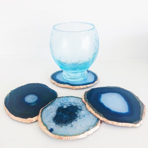 May include: A collection of blue agate coasters with gold edges, alongside a matching blue glass. The coasters vary in shape and pattern, with a polished finish. The glass has a textured design and sits on a coaster.