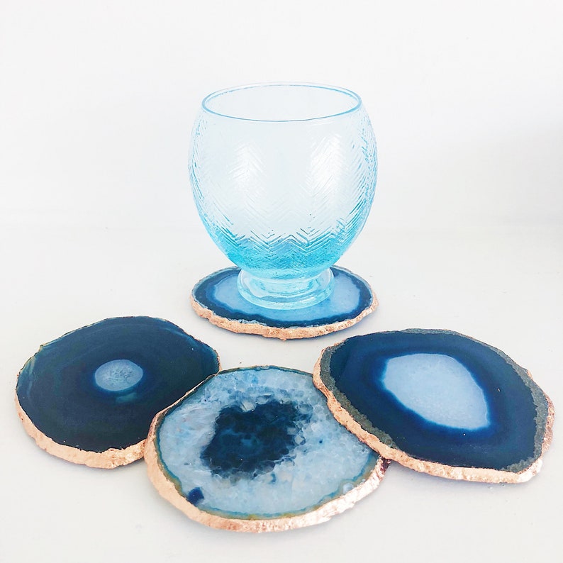 May include: A collection of blue agate coasters with gold edges, alongside a matching blue glass. The coasters vary in shape and pattern, with a polished finish. The glass has a textured design and sits on a coaster.