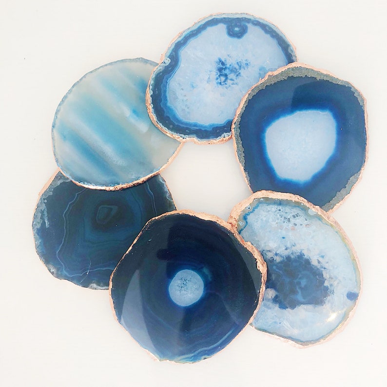 May include: A collection of seven blue agate coasters with copper-colored edges. The coasters vary in shades of blue, from light to deep indigo, and are arranged in a circular pattern against a white background. These decorative coasters add a touch of elegance to any home.