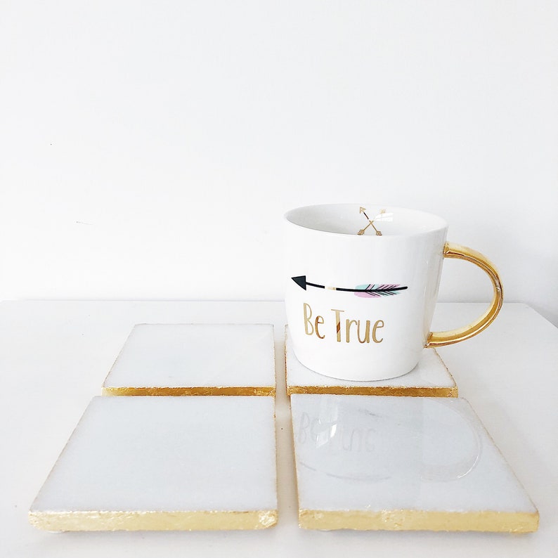 May include: Four white square coasters with gold trim and a white mug with a gold handle. The mug has a black and pink arrow with the text "Be True" in gold lettering.