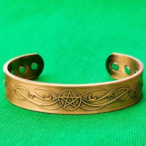 以下が含まれることがあります： 五芒星と渦巻き模様が刻まれたブロンズ色のカフブレスレット。 オープンエンドのブレスレットは装飾的なデザインで、緑色の背景に配置されています。 スピリチュアルまたはウィッカをテーマにしたアクセサリーです。