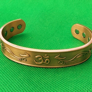 Puede incluir: Una pulsera de brazalete dorada con un símbolo Om grabado y un diseño floral. La pulsera abierta tiene tres agujeros circulares en cada extremo y se muestra sobre una superficie verde.