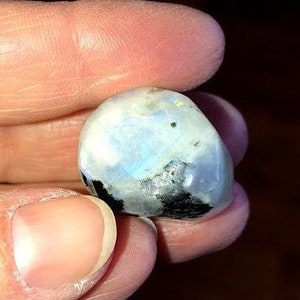 May include: A smooth, white, and iridescent moonstone with black inclusions. The stone is held in a hand.