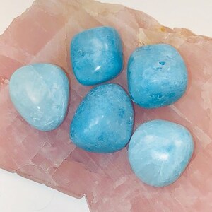 May include: Five smooth, light blue tumbled stones on a pink, textured surface. The stones are various sizes and shapes.