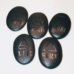 May include: Five black oval-shaped stones with different symbols and words engraved on them. The words are "EARTH", "SPIRIT", "WATER", "AIR", and "FIRE".