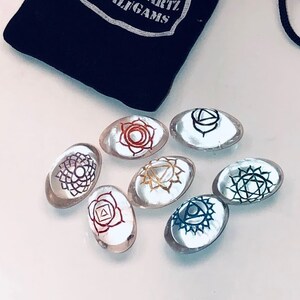 Może przedstawiać: Siedem przezroczystych, owalnych kamieni z symbolami czakr w r&oacute;żnych kolorach. Kamienie są ułożone w k&oacute;łko na białym tle. Kamienie znajdują się w czarnym woreczku ze sznurkiem, na kt&oacute;rym nadrukowany jest tekst "Chakra Quartz Shivaligams".