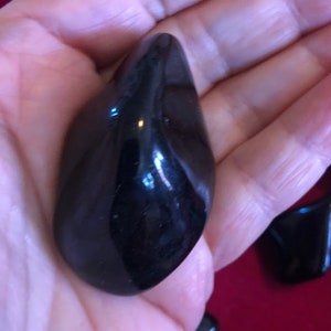 May include: A smooth, black, polished stone with a rounded, teardrop shape. The stone is held in a hand, and the background is a red surface.