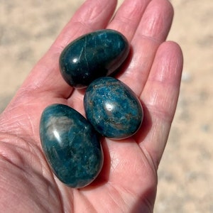 Puede incluir: Tres huevos de cristal de apatita azul verdoso, pulidos y redondeados, en una mano.
